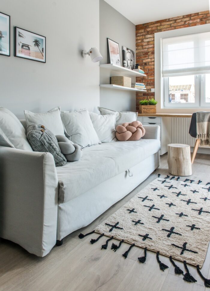 Sofa w pokoju gościnnym wydziela strefę wypoczynkową. Ściana za nią jest pomalowana jasną szarością. Sofa jest...
