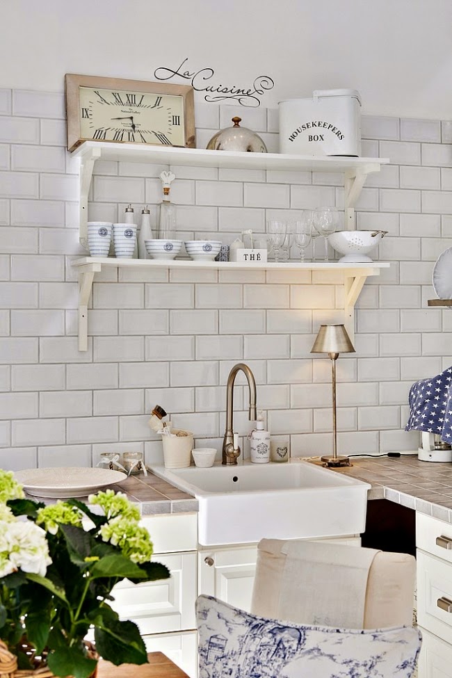 biała płytka glazurowana cegiełka na scianie w kuchni,płytka glazurowana cegiełka,ceramiczna cegiełka w białym kolorze,białe stylowe półki z drewna,białe półki do kuchni,ceramiczny skandynawski zlewozmywak,biała kuchnia w stylu skandynawskim,skandynawsko-prowansalska mieszanka w aranzacji kuchennej,aranzacja białej kuchni w mieszanym stylu scandy i provence,metalowe białe puszki z napisami,kuchenne metalowe pojemniki,emaliowane puszki i pudełka na przechowwyanie produktów w kuchni,biała stylowa galenteria z porcelany,kuchenne drobiazgi w prowansalskim stylu,szklane słoiki i pojemniki,ozdoby w stylu shabby chic w białej kuchni,tekstylia do kuchni