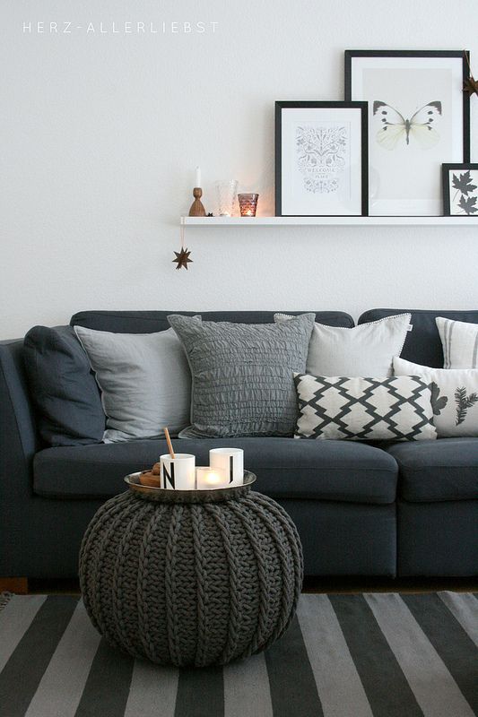 Ciemna sofa w salonie udekorowana dużą ilością poduszek w szarym kolorze. Nad sofą biała wąska półeczka na której...