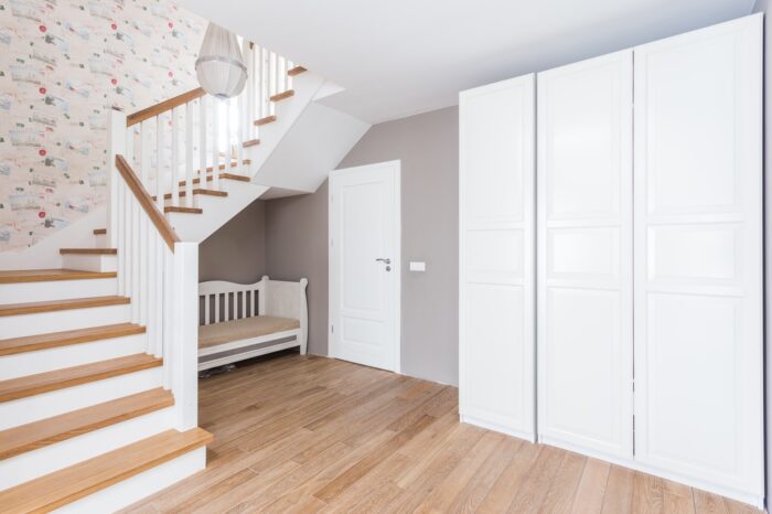 Obszerny hol ze schodami na górę. Biała duża szafa i ławeczka pod schodami idealnie współgrają z drewnianą podłogą i...